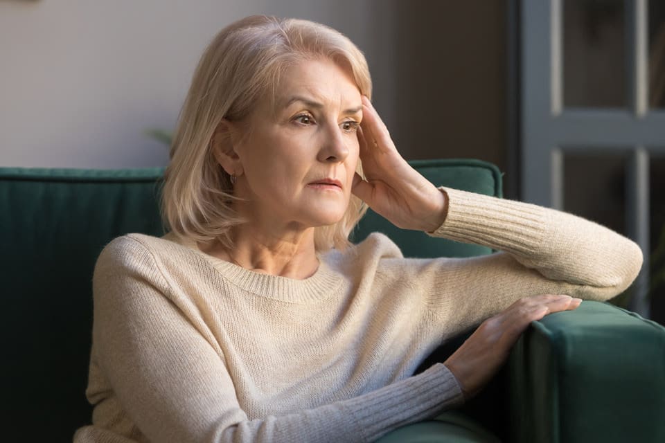 A senior woman sits on the couch while resting her head against her hand.